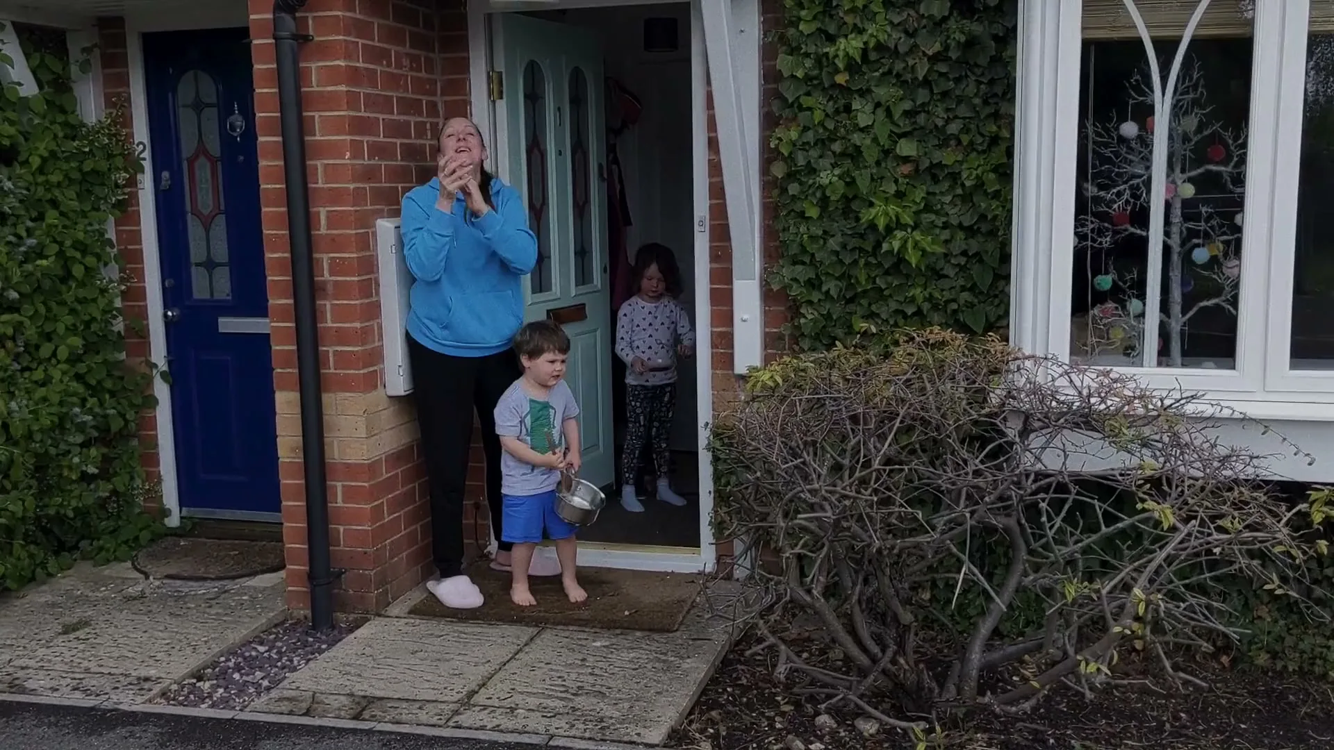 A woman and two children stand in their doorway, clapping and making noise with pots and pans.