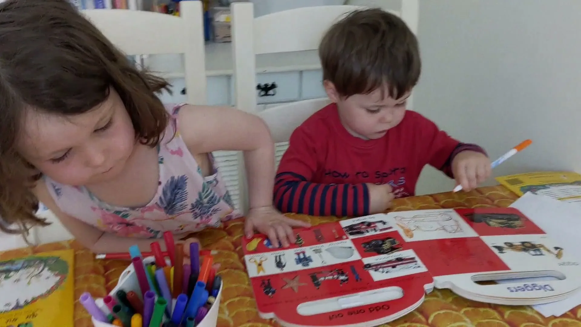Two young children, a girl and a boy, are sitting at a table, coloring in a book with markers and pencils.