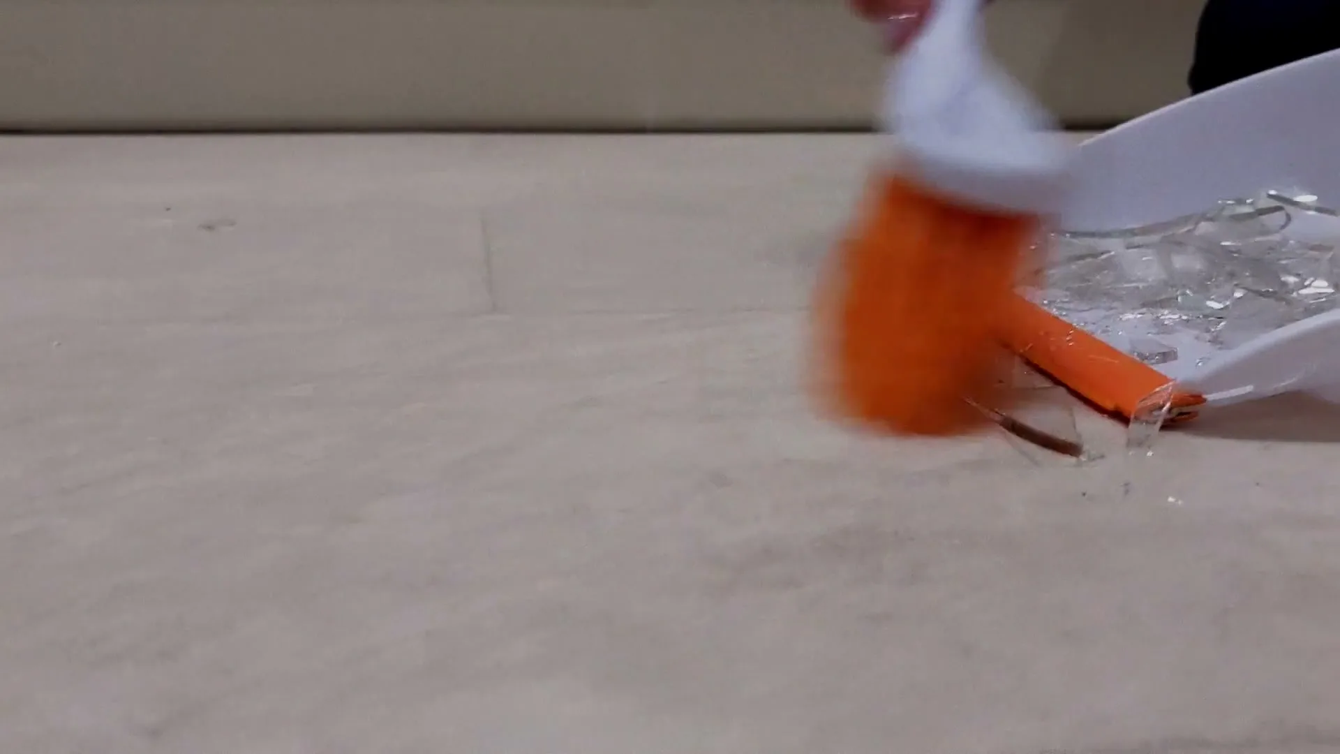 A person sweeps broken glass into a dustpan with an orange brush on a light-colored floor, suggesting a domestic accident or violence aftermath.