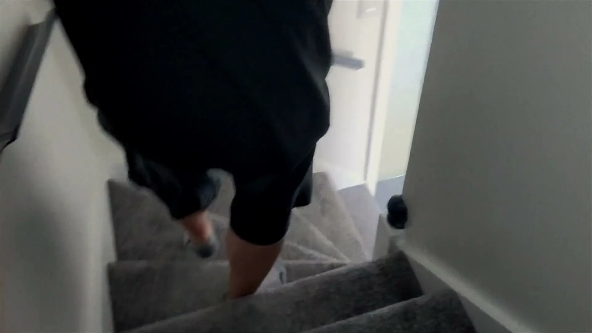 A person in black shorts and a dark t-shirt walks down a carpeted staircase in a house, seen from a high angle.