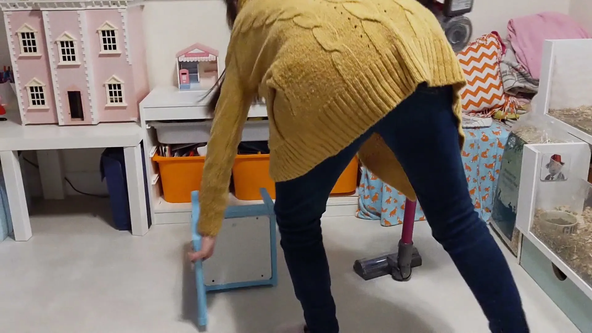 A person in a yellow sweater and blue jeans vacuums a child's room, moving a small blue chair out of the way.