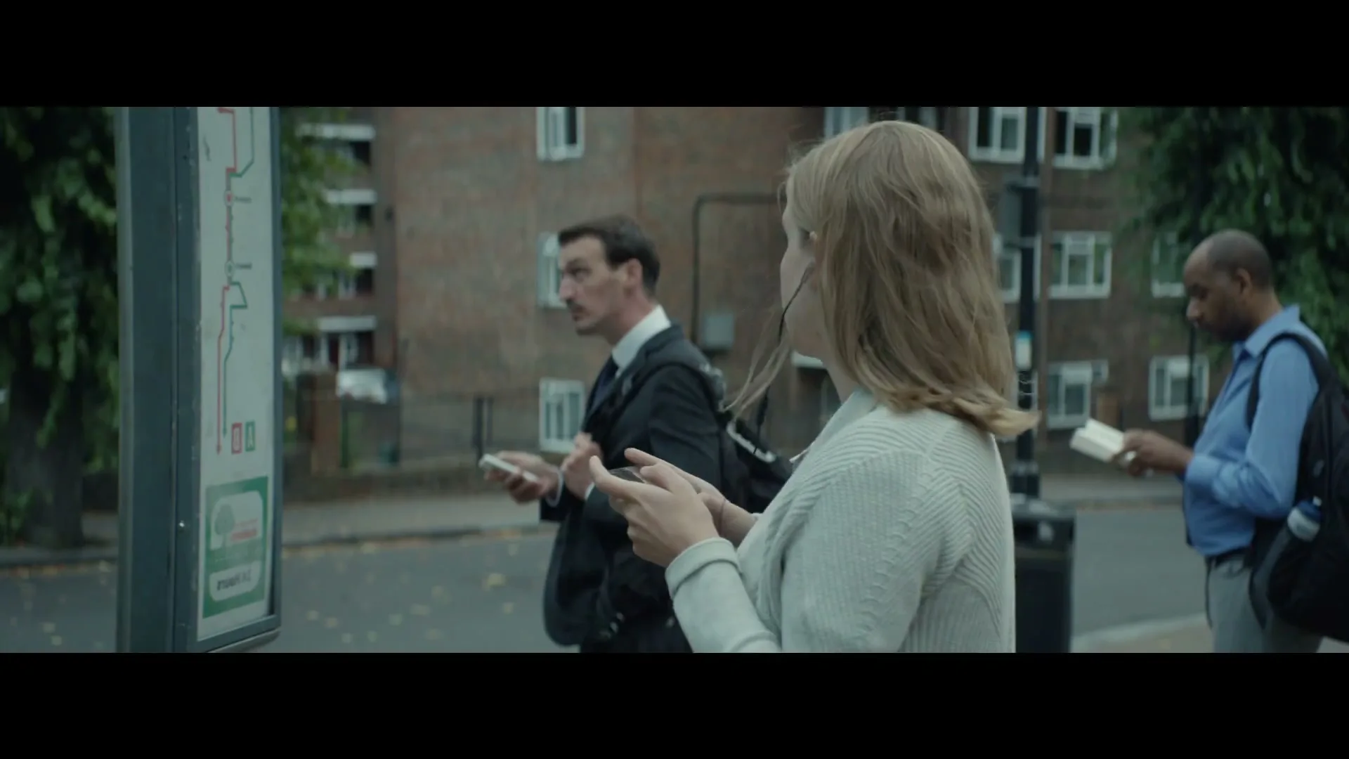 A woman with blonde hair and earbuds looks up from her phone while waiting at a bus stop with other people.