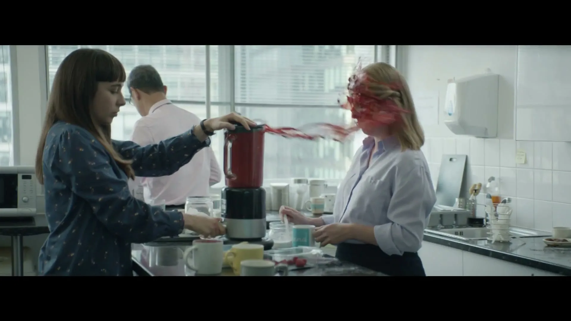 A woman in a blue shirt accidentally sprays red liquid from a blender onto a blonde woman in a white shirt in an office kitchen.