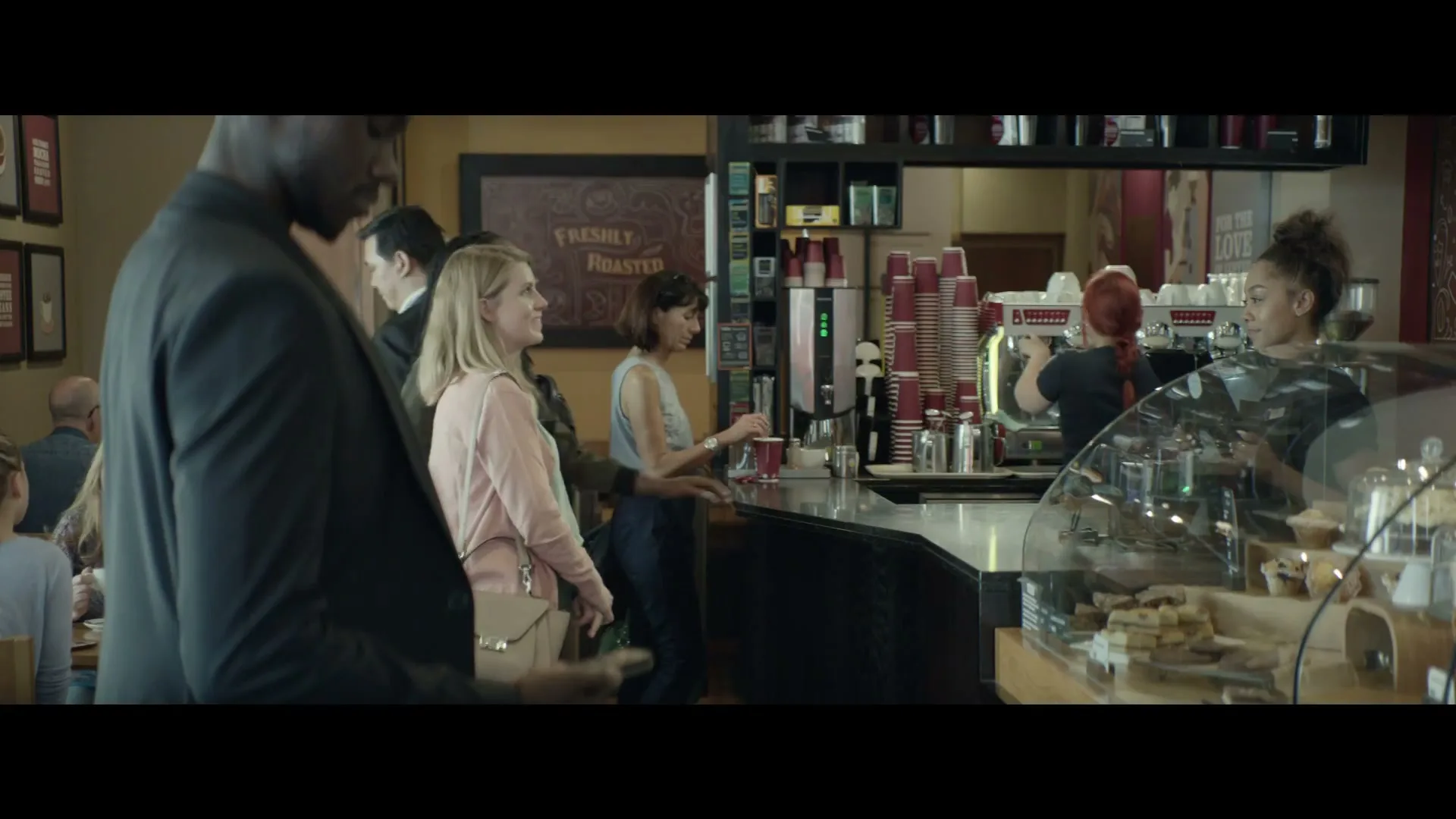 A diverse group of people wait in line at a coffee shop, with baristas working behind the counter.