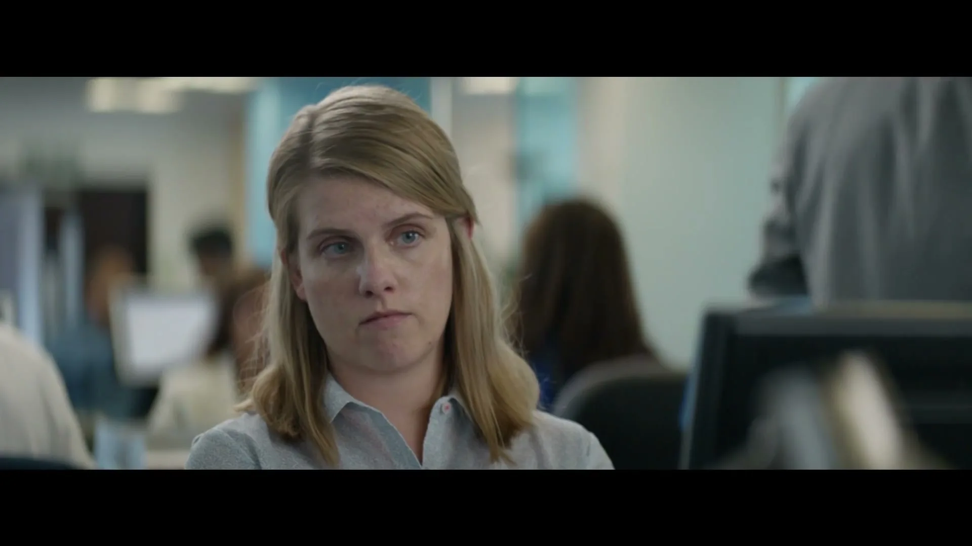A young woman with blonde hair, wearing a light blue shirt, sits at a desk in an office, looking around with a neutral expression.