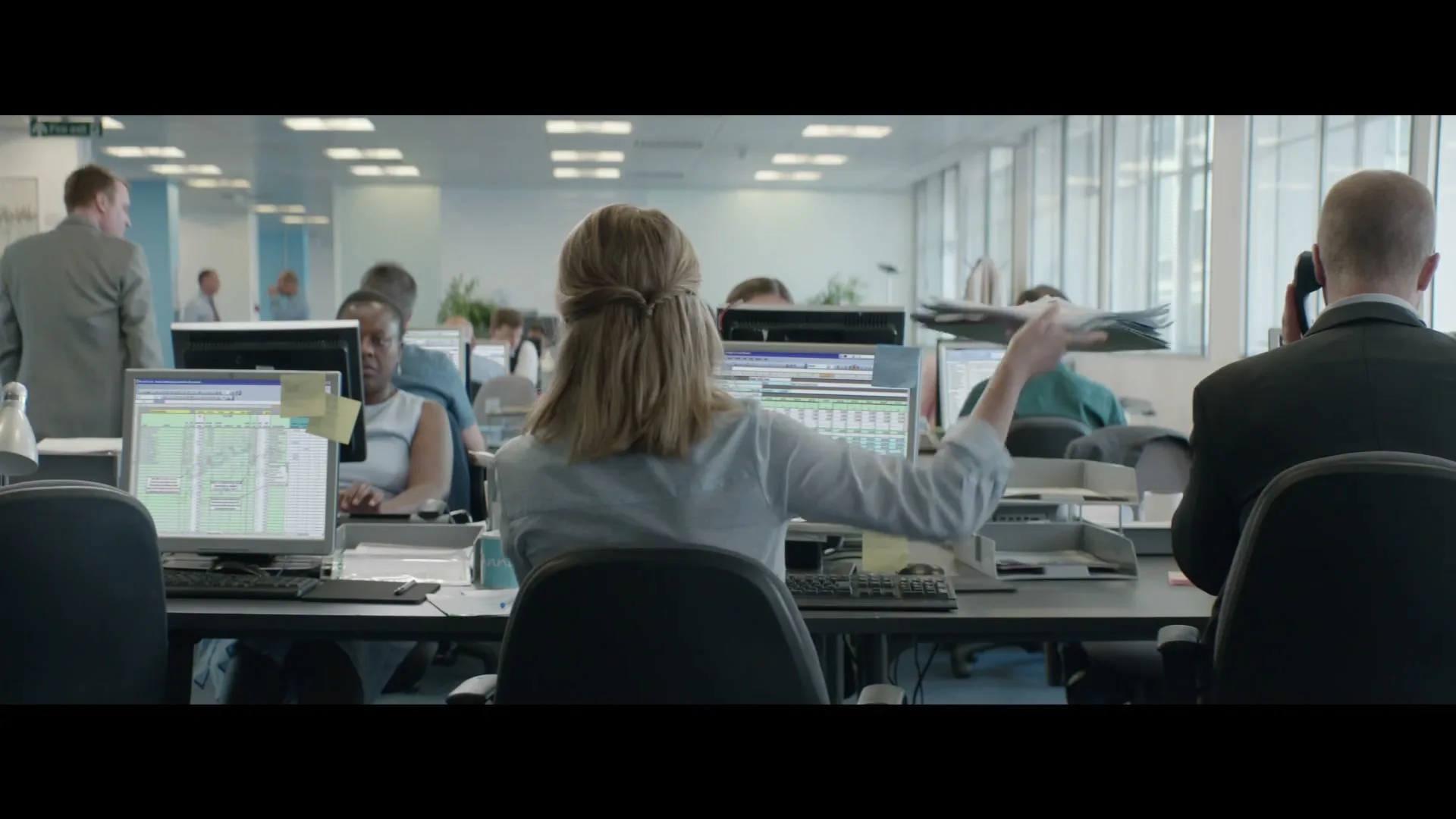 A woman with blonde hair, seen from behind, sits at a desk in a busy open-plan office, waving her hands in frustration.