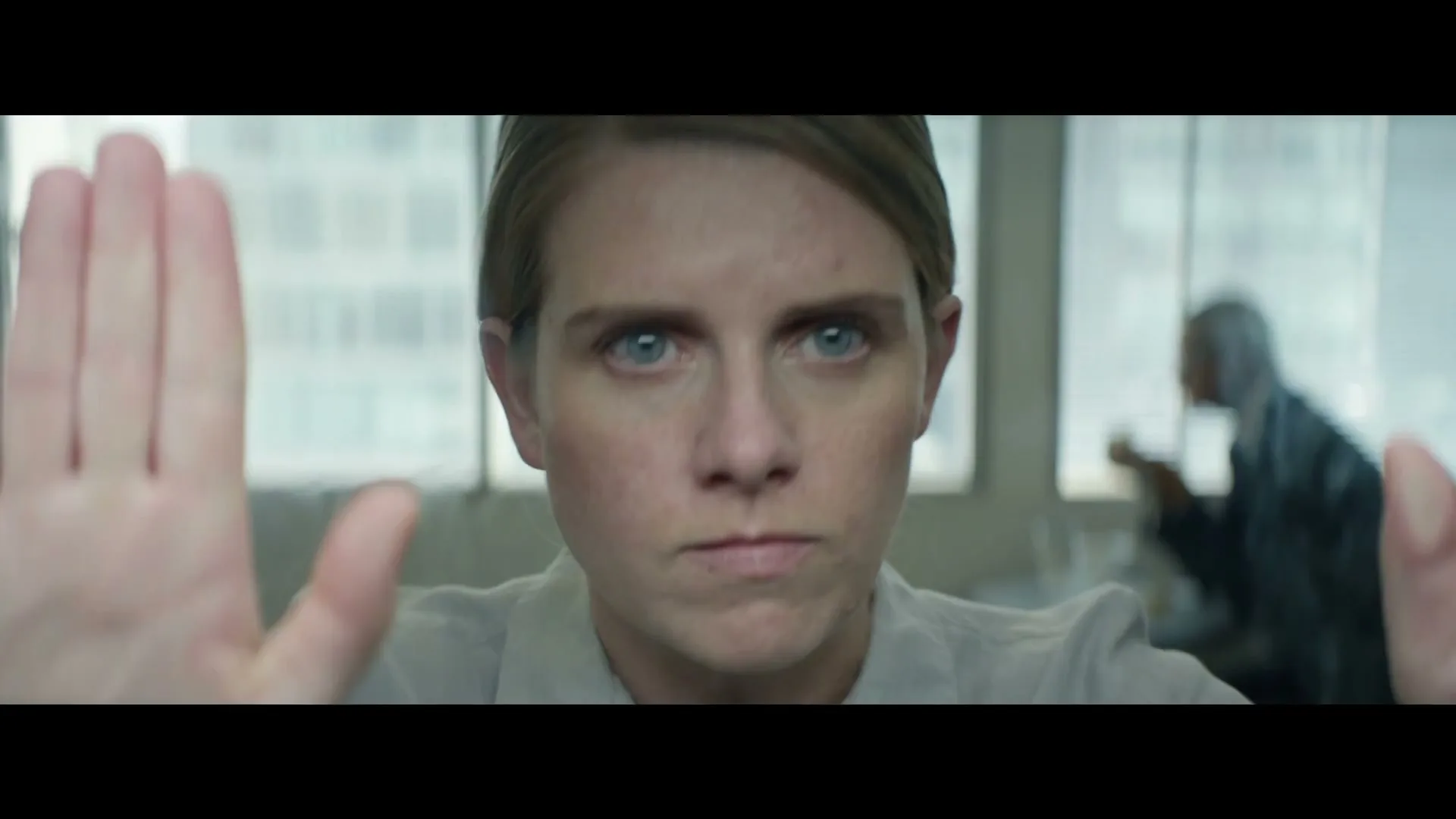 A woman with blonde hair and blue eyes stares intensely at the camera, holding her hands up as if to stop something, in an office setting.
