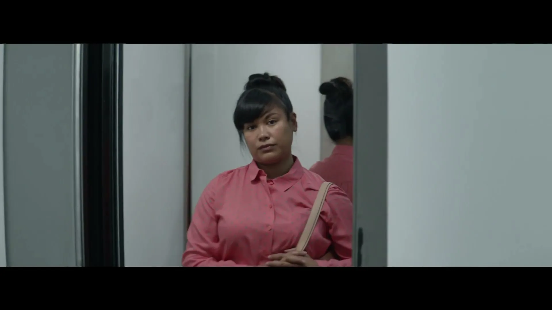 A young woman in a pink shirt stands in a hallway, looking thoughtful, with her reflection visible in a mirror.
