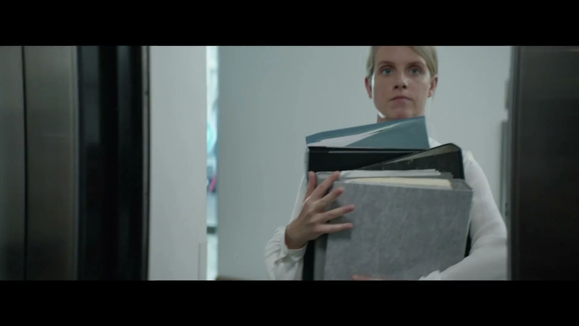 A woman in a white shirt, carrying a stack of folders, exits an elevator in an office building.