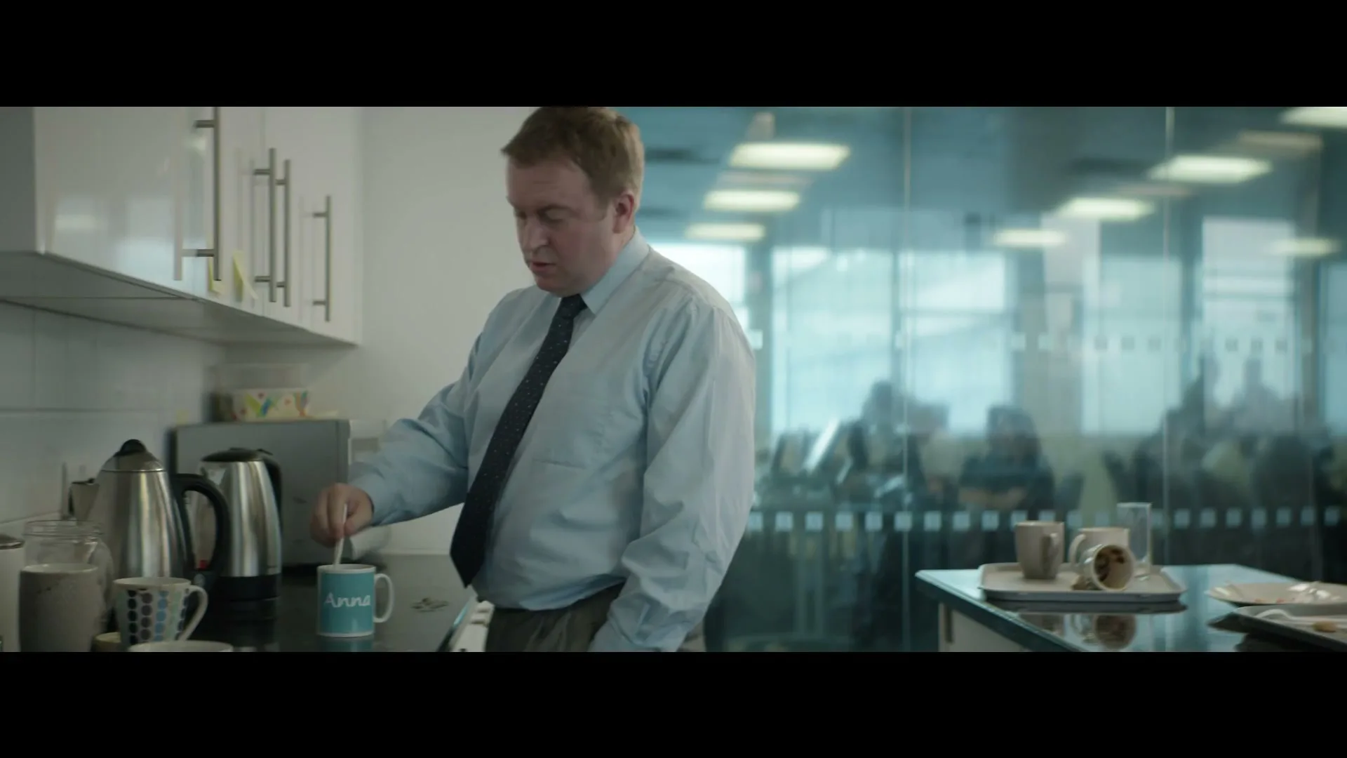 A man in a light blue shirt and dark tie stirs a mug in an office kitchen, looking around with a slightly concerned expression.