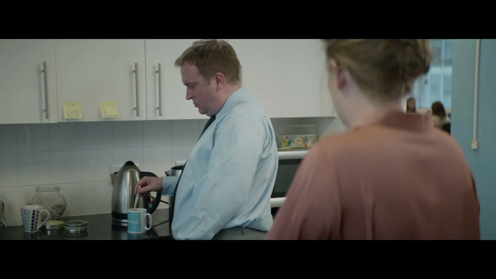 A man in a light blue shirt and tie looks over his shoulder at a woman in a peach top in an office kitchen.