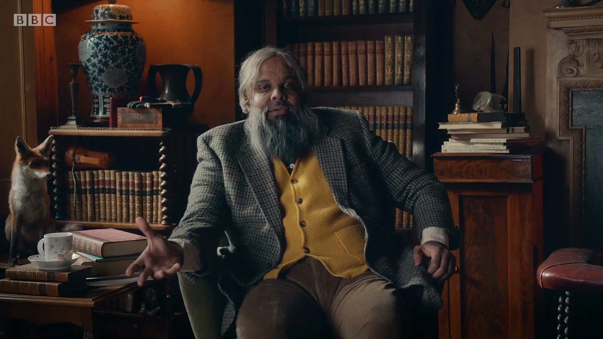 A middle-aged man with a long grey beard and tweed jacket sits in a plush armchair, gesturing while speaking in a dimly lit, wood-paneled study.