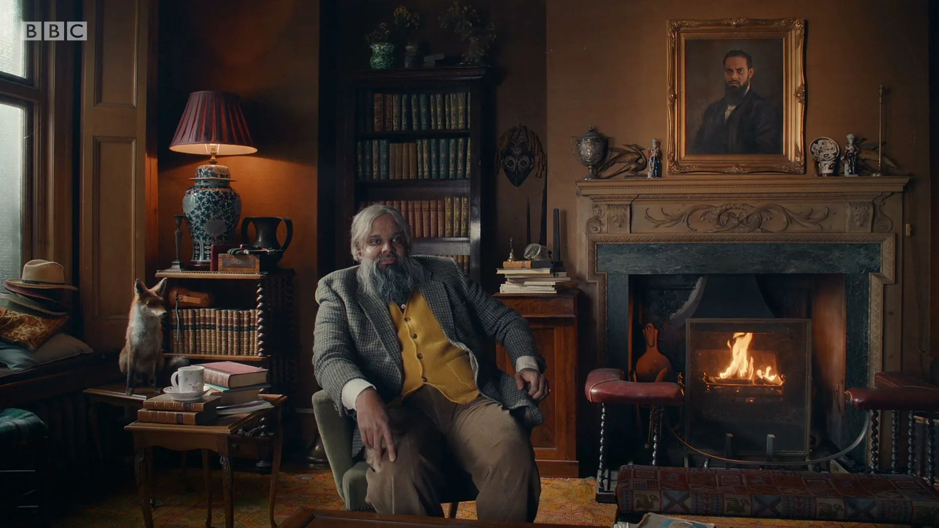 A man with a long grey beard and a yellow vest sits in an armchair in a cozy, book-filled study with a fireplace.