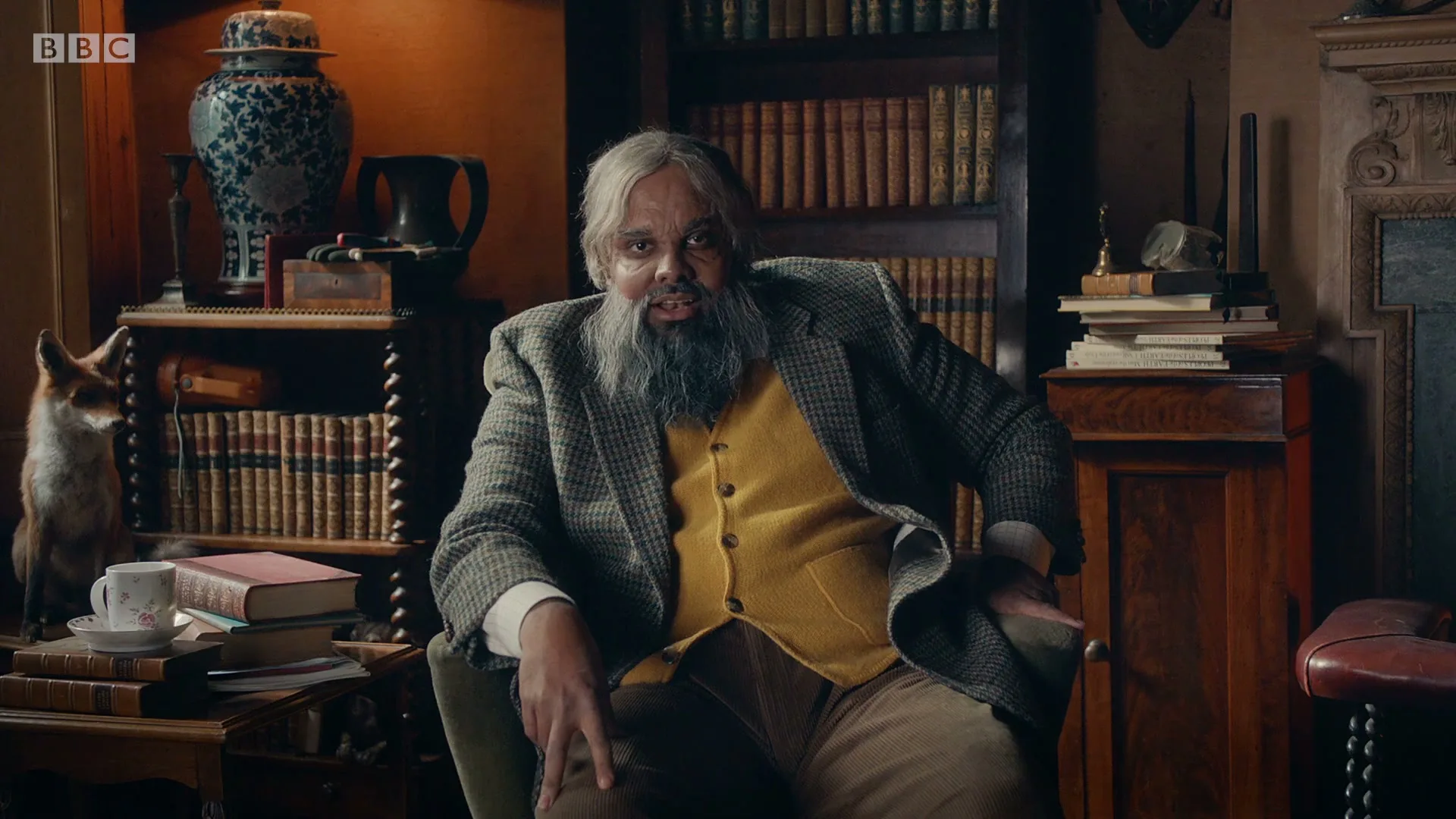 A man with a long grey beard and a yellow vest sits in a chair in a dimly lit, old-fashioned study, speaking directly to the camera.