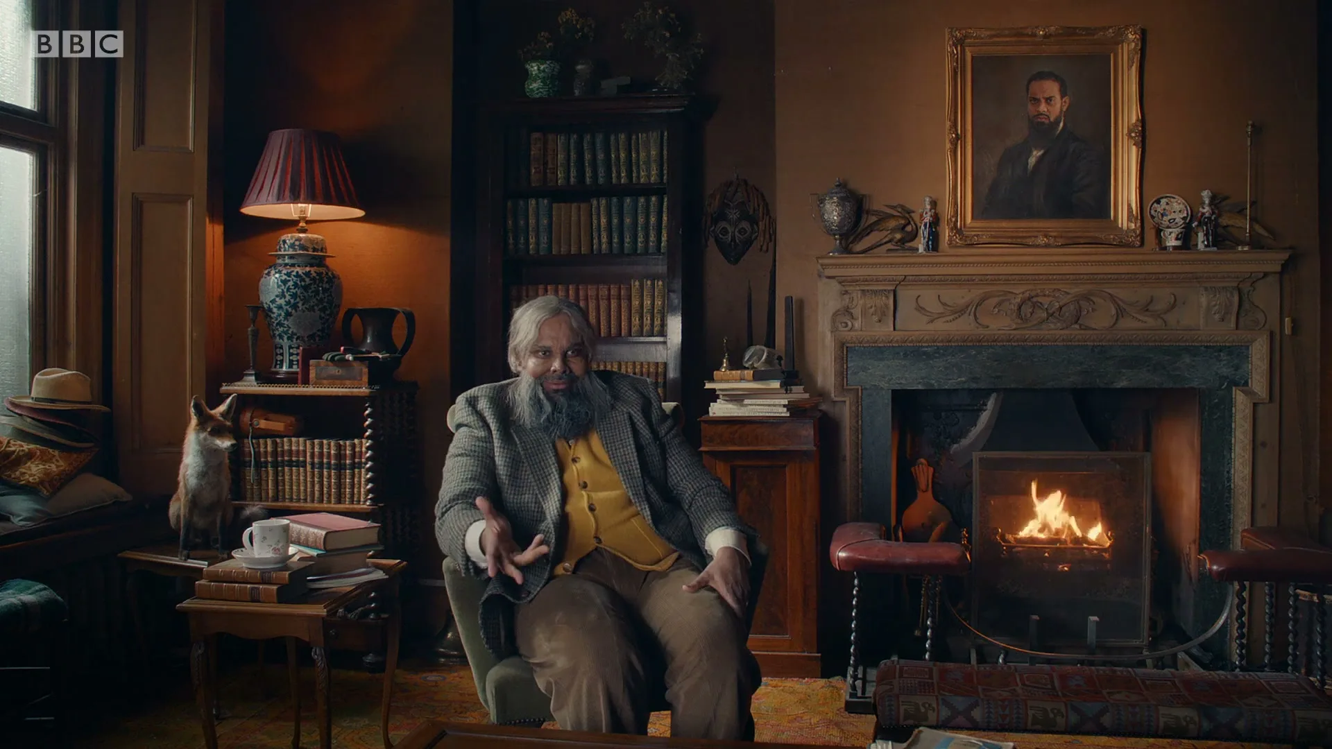 A man with a long white beard and grey hair sits in an armchair in a cozy, book-filled room with a fireplace, gesturing as he speaks.