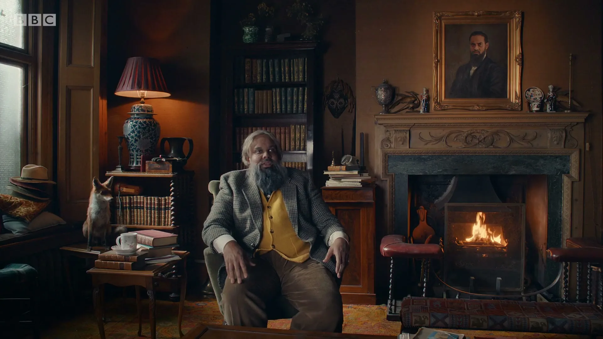 A bearded man in a tweed jacket and yellow vest sits in an armchair in a dimly lit, ornate study with a fireplace and bookshelves.