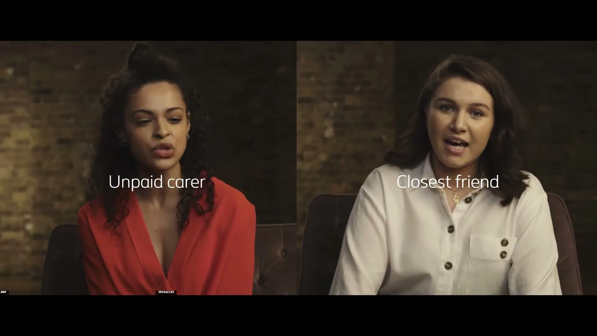 Two women, one with curly hair in a red top and the other with dark hair in a white shirt, sit side-by-side on a couch, talking to the camera.