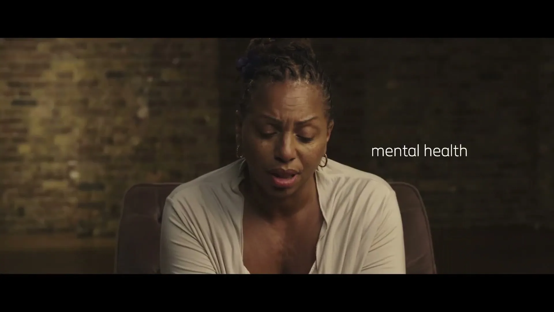 A Black woman with dreadlocks, wearing a light cardigan, sits in a chair with a sad expression as text appears on screen in a dimly lit room.