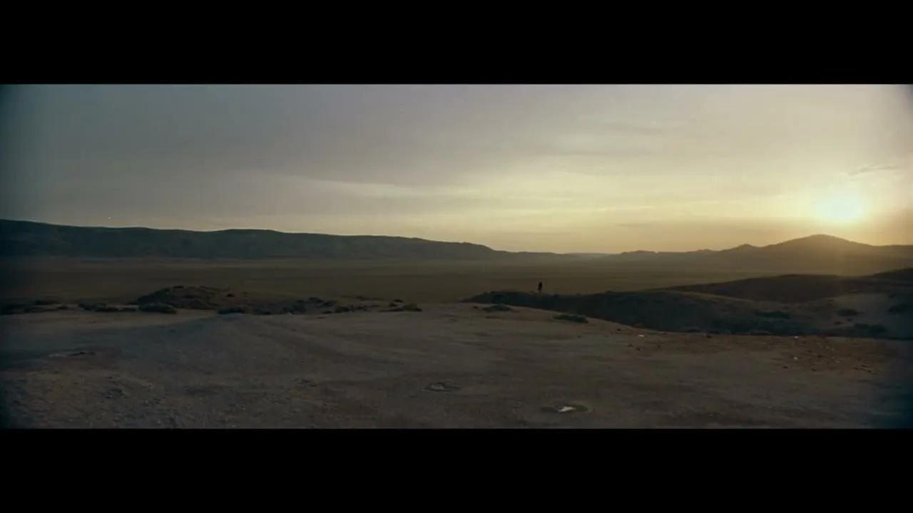A lone figure stands on a hill overlooking a vast desert landscape with mountains in the distance under a dramatic sky at sunset.