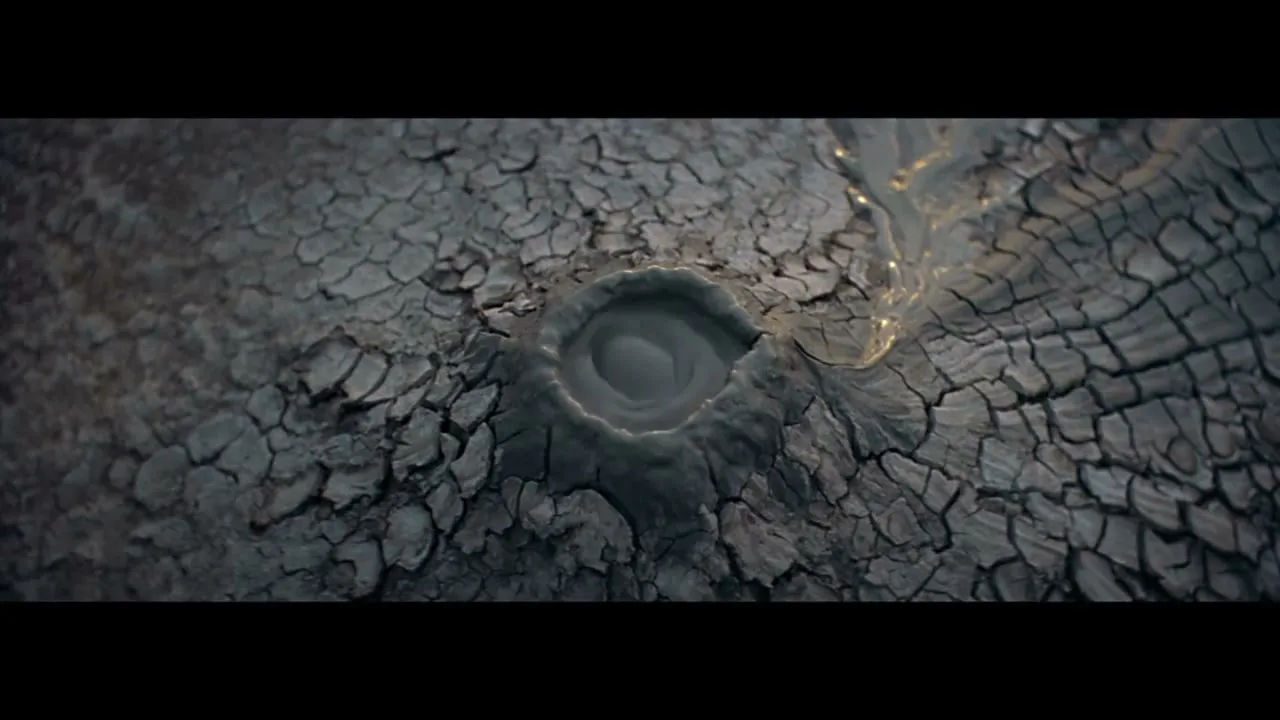 A wide shot shows a small pool of water in the center of a cracked, dry, dark gray landscape, with a golden liquid flowing into it from the right.