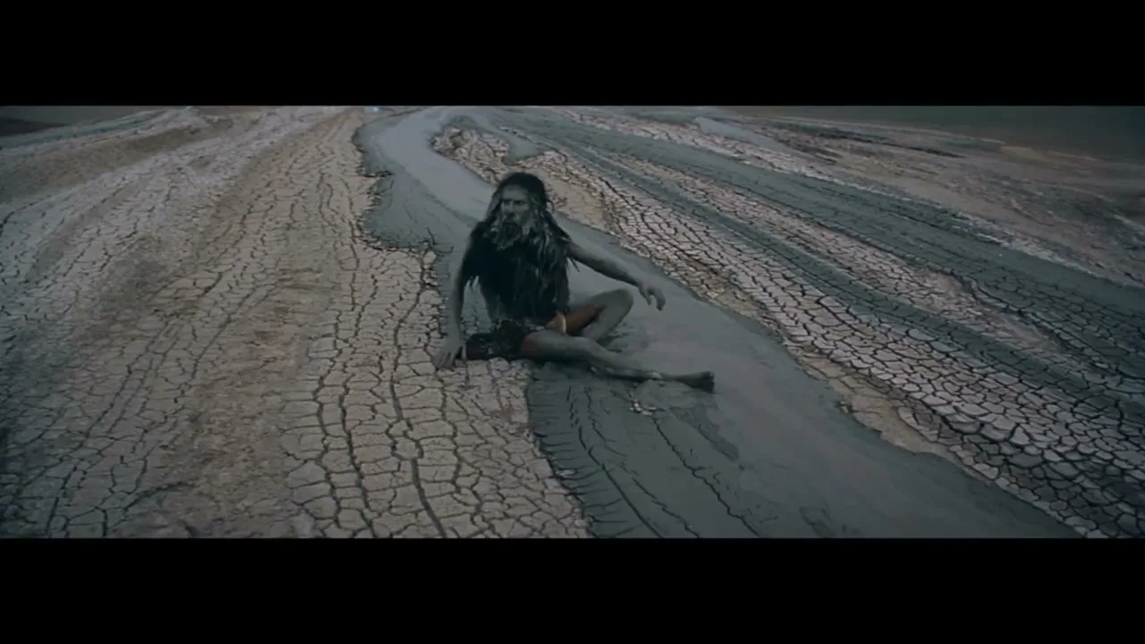 A person with long dark hair and mud-covered skin sits in a muddy riverbed, surrounded by cracked earth, looking around.