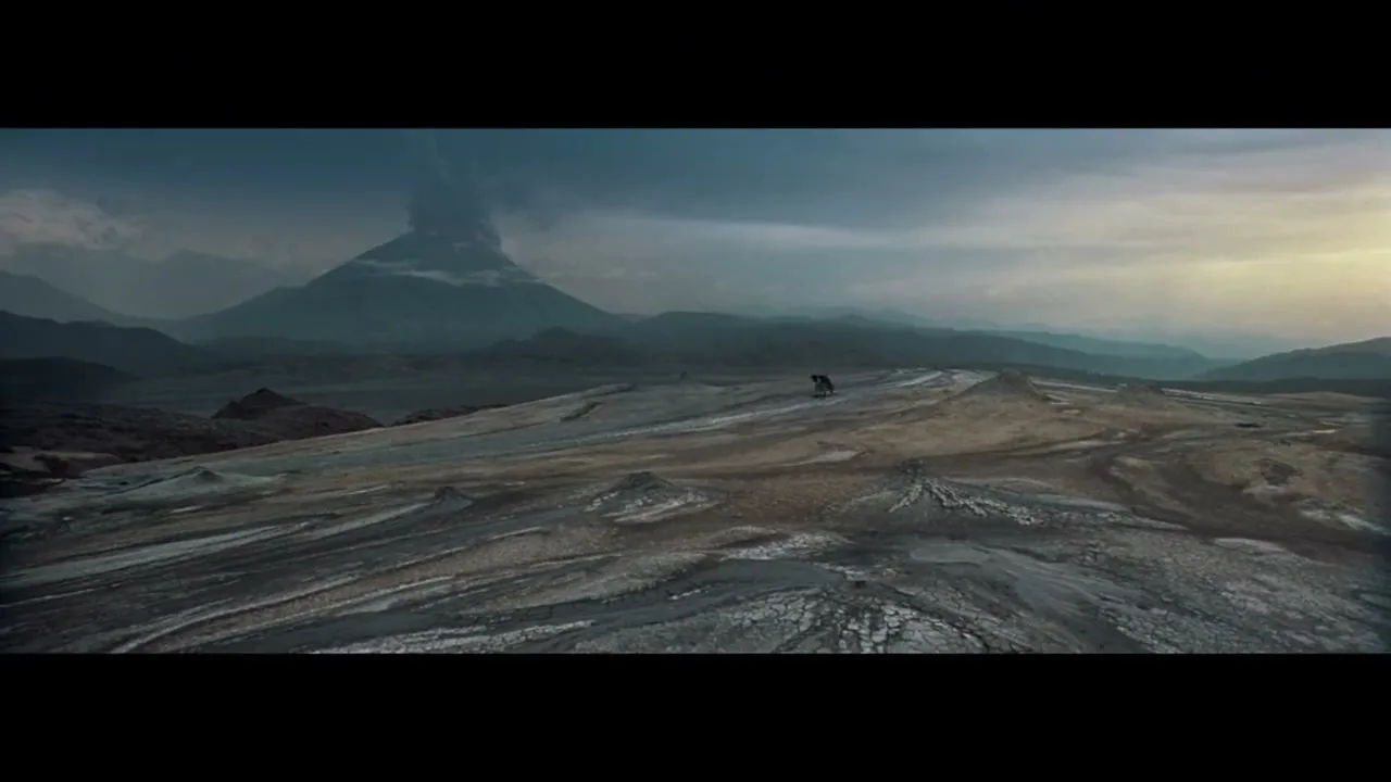 A lone figure stands on a desolate, rocky landscape as a volcano erupts in the distance under a dramatic sky.
