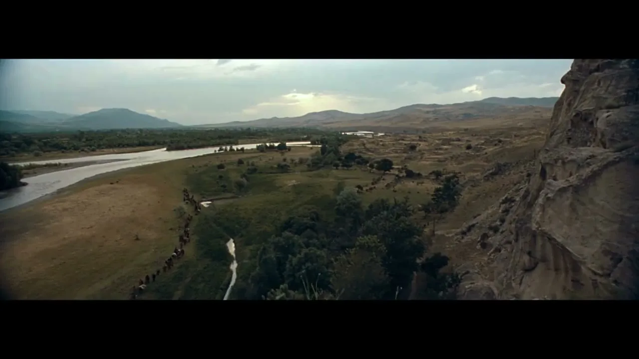 A wide shot shows a long line of people on horseback riding along a river in a vast, mountainous landscape under a cloudy sky.