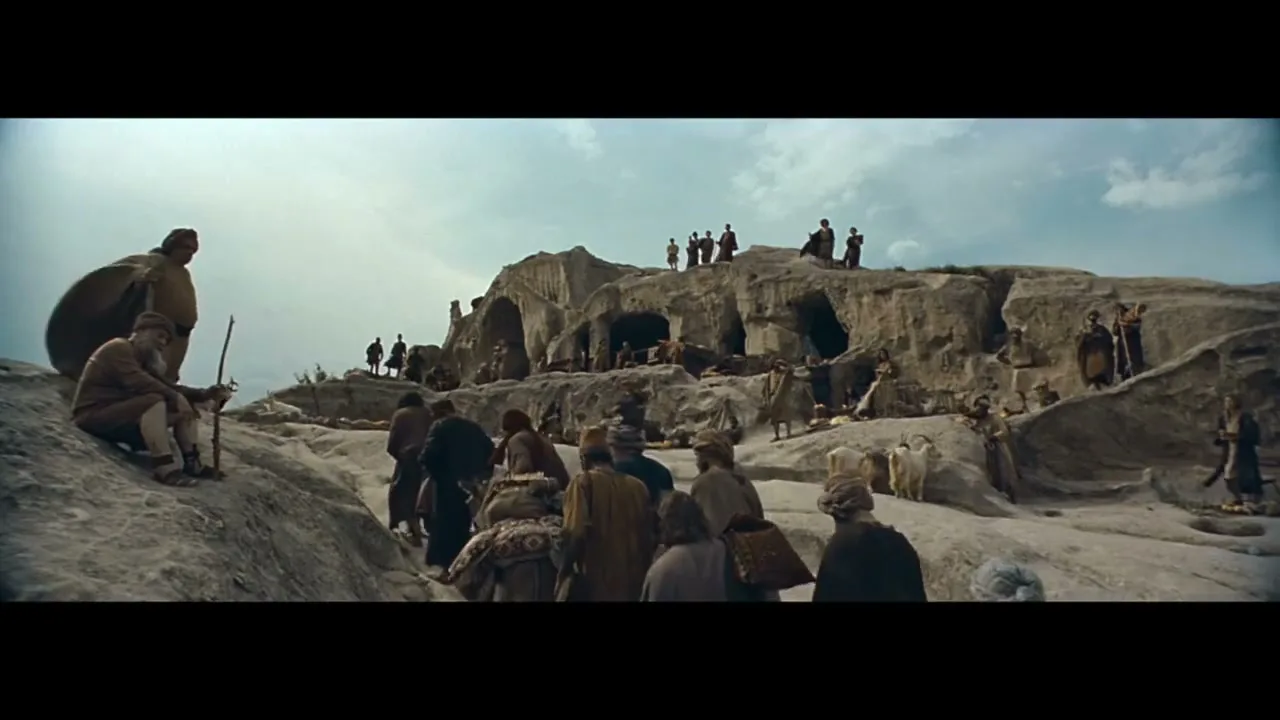 A wide shot shows a large group of people in ancient attire walking through a rocky, mountainous landscape under a cloudy sky.