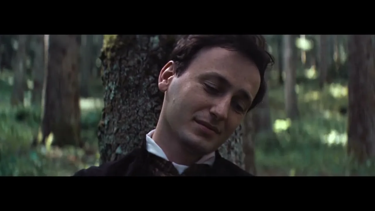 A man with dark hair and a white shirt leans against a tree in a forest, looking content.
