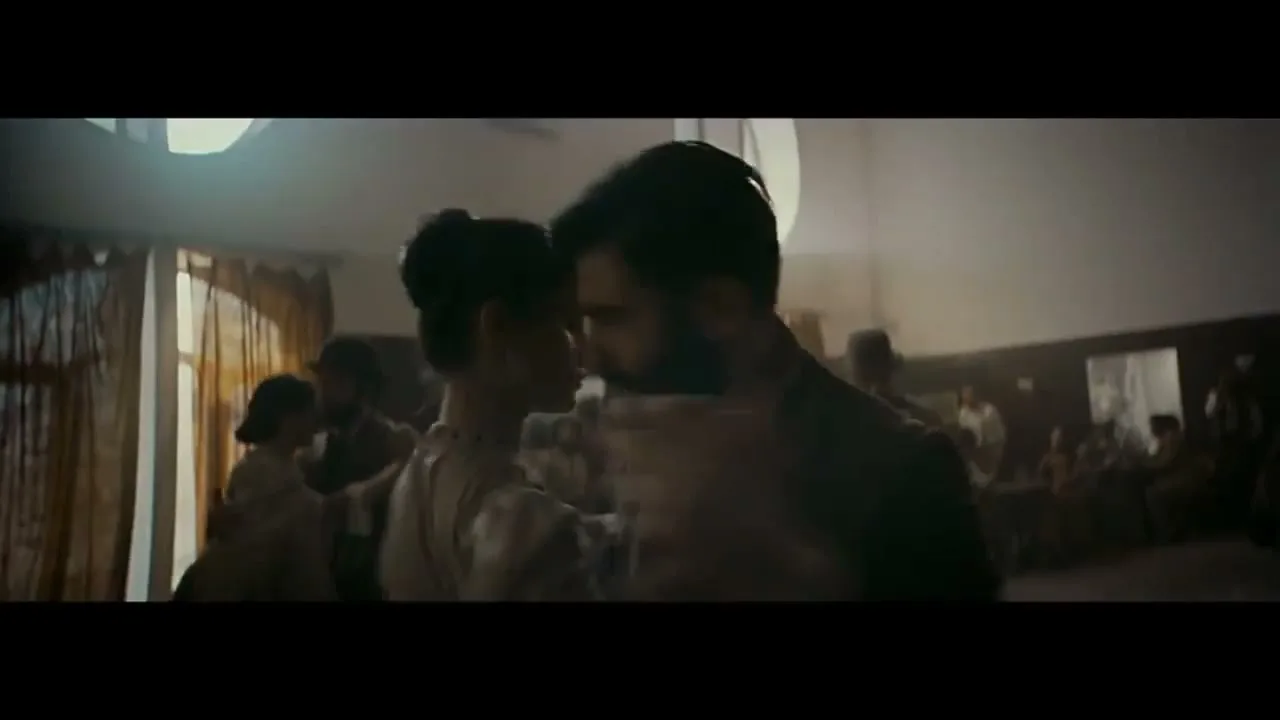 A young couple, dressed in period clothing, dances closely in a dimly lit room with other people in the background.