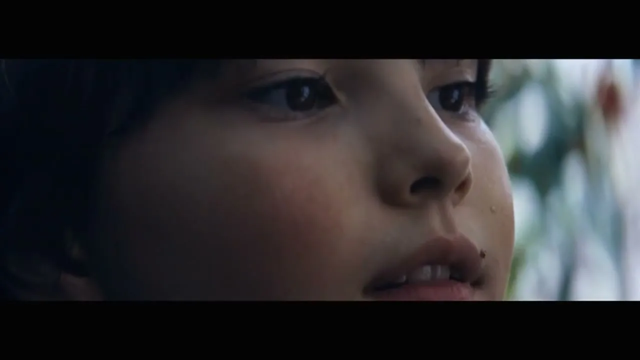 A young child with dark hair and a small cut on their lip looks up with a curious expression, with soft light on their face.