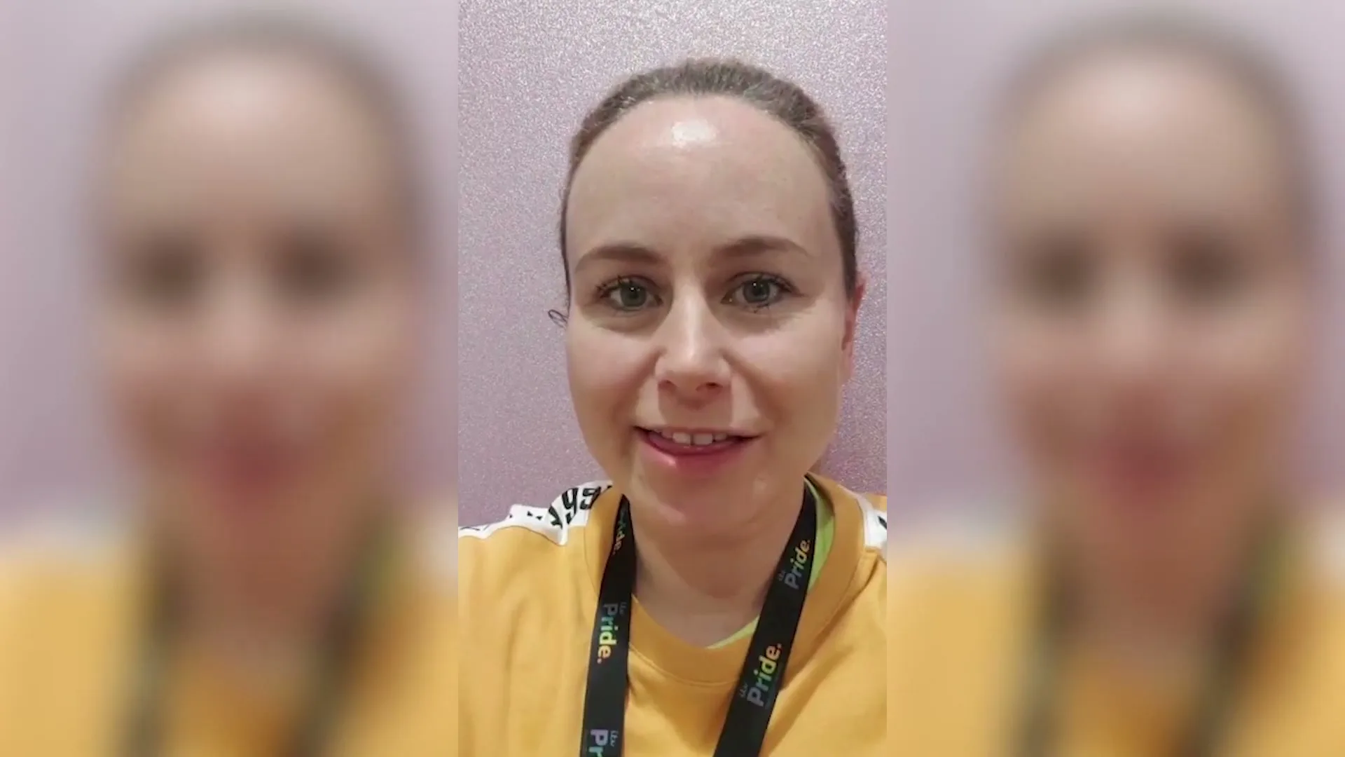 A woman with light skin and hair pulled back smiles and talks directly into the camera, wearing a yellow top and a lanyard.