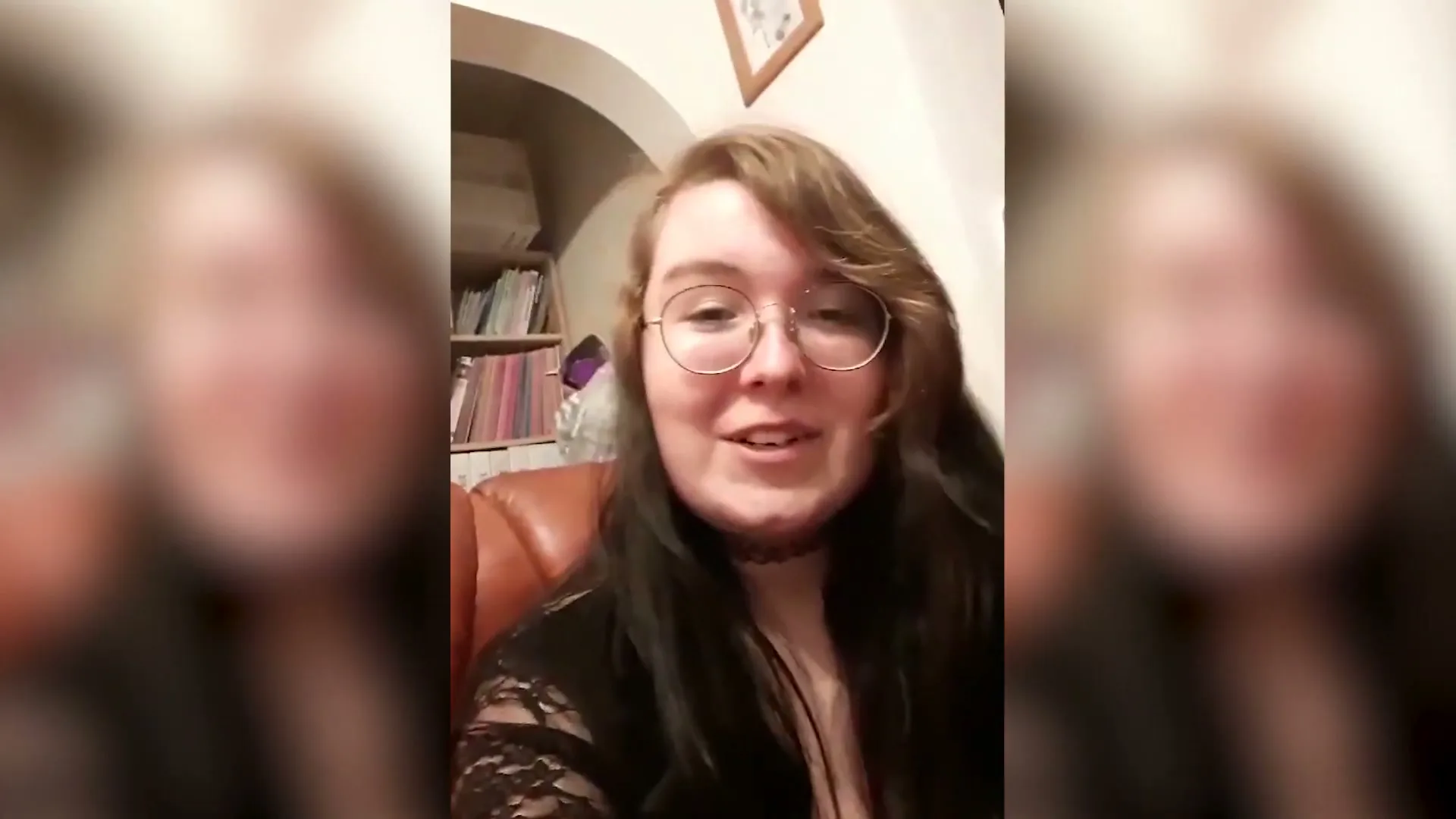 A young woman with glasses and long dark hair, wearing a black top, talks to the camera in a cozy living room at night.