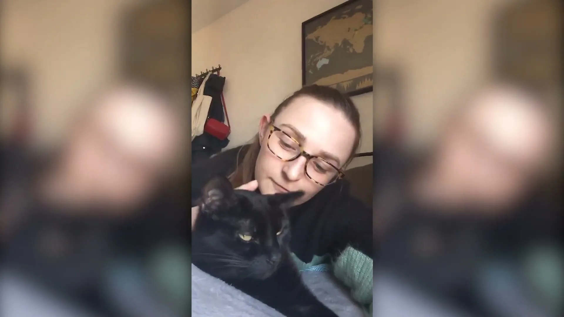 A young woman with glasses pets a black cat while lying on a bed, filmed from a phone's perspective in a cozy room.
