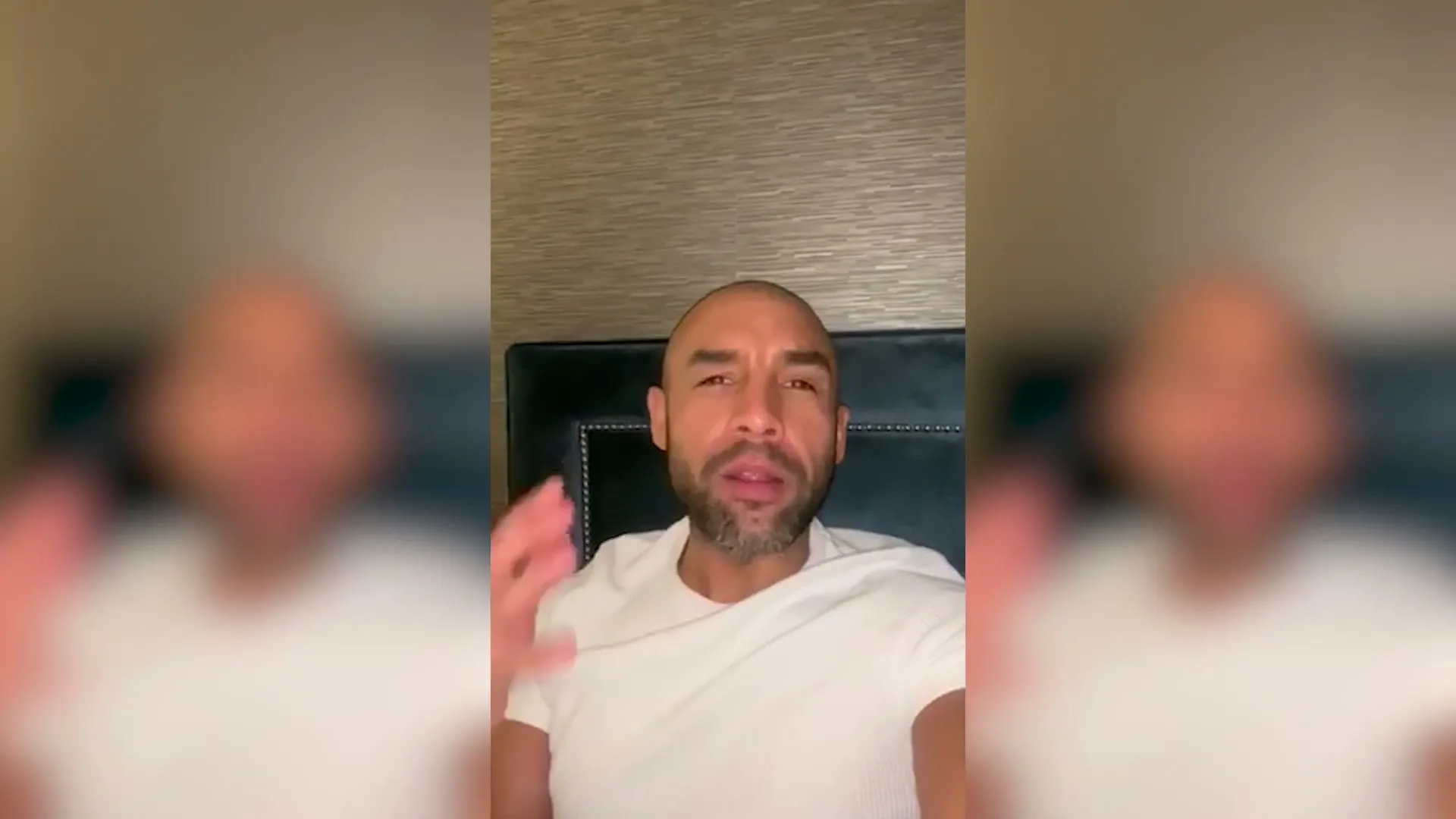 A bald man in his 30s, wearing a white t-shirt, talks directly to the camera from his bed in a bedroom.