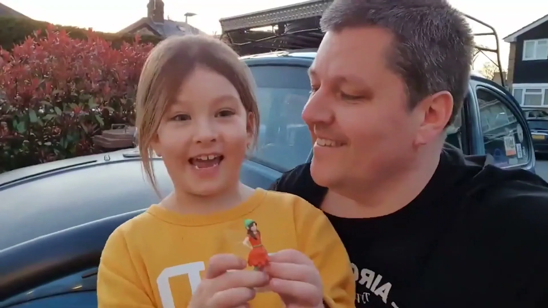A young girl in a yellow top holds a small fairy doll, smiling at the camera next to a man in a black t-shirt, outdoors by a car.