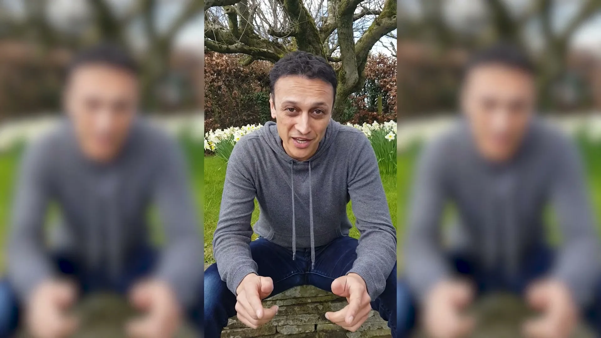 A man in his 30s, wearing a grey hoodie and jeans, crouches outdoors in a garden with daffodils, speaking directly to the camera.