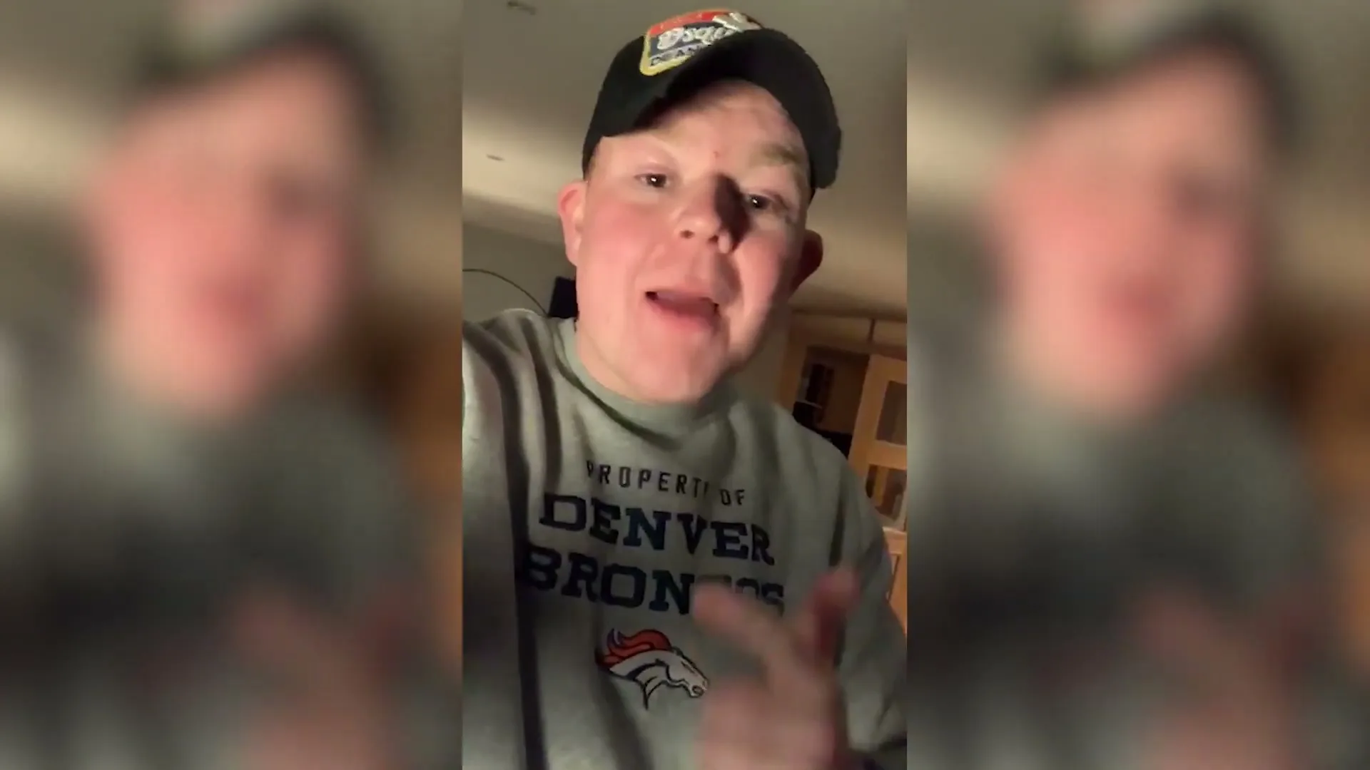 A young man in a Denver Broncos sweatshirt and baseball cap talks directly to the camera, gesturing with his hands in an interior setting.