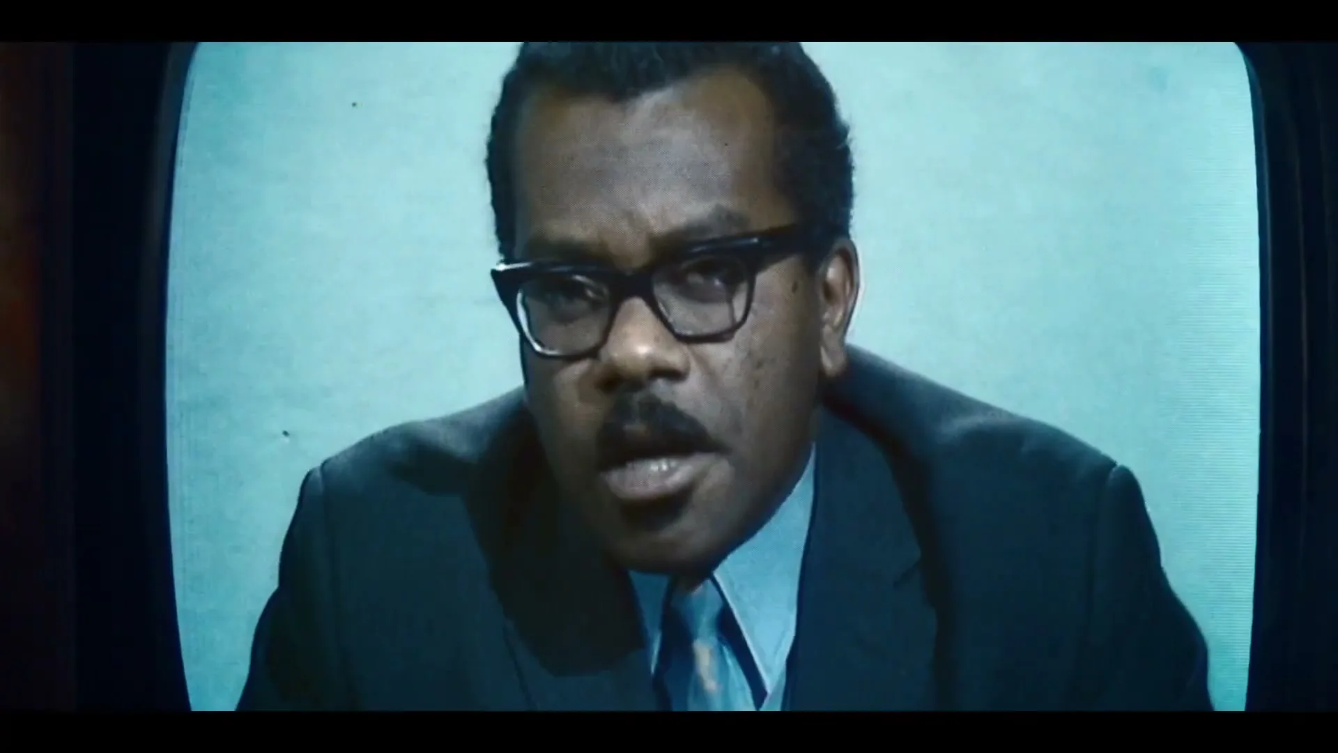 A man with dark skin, glasses, and a mustache, wearing a suit and tie, speaks directly to the camera on a vintage television screen with static interference.