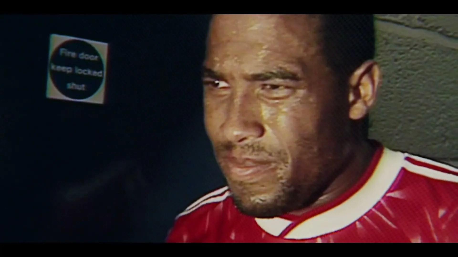 A sweaty male athlete in a red and white jersey looks distressed in a dimly lit locker room.