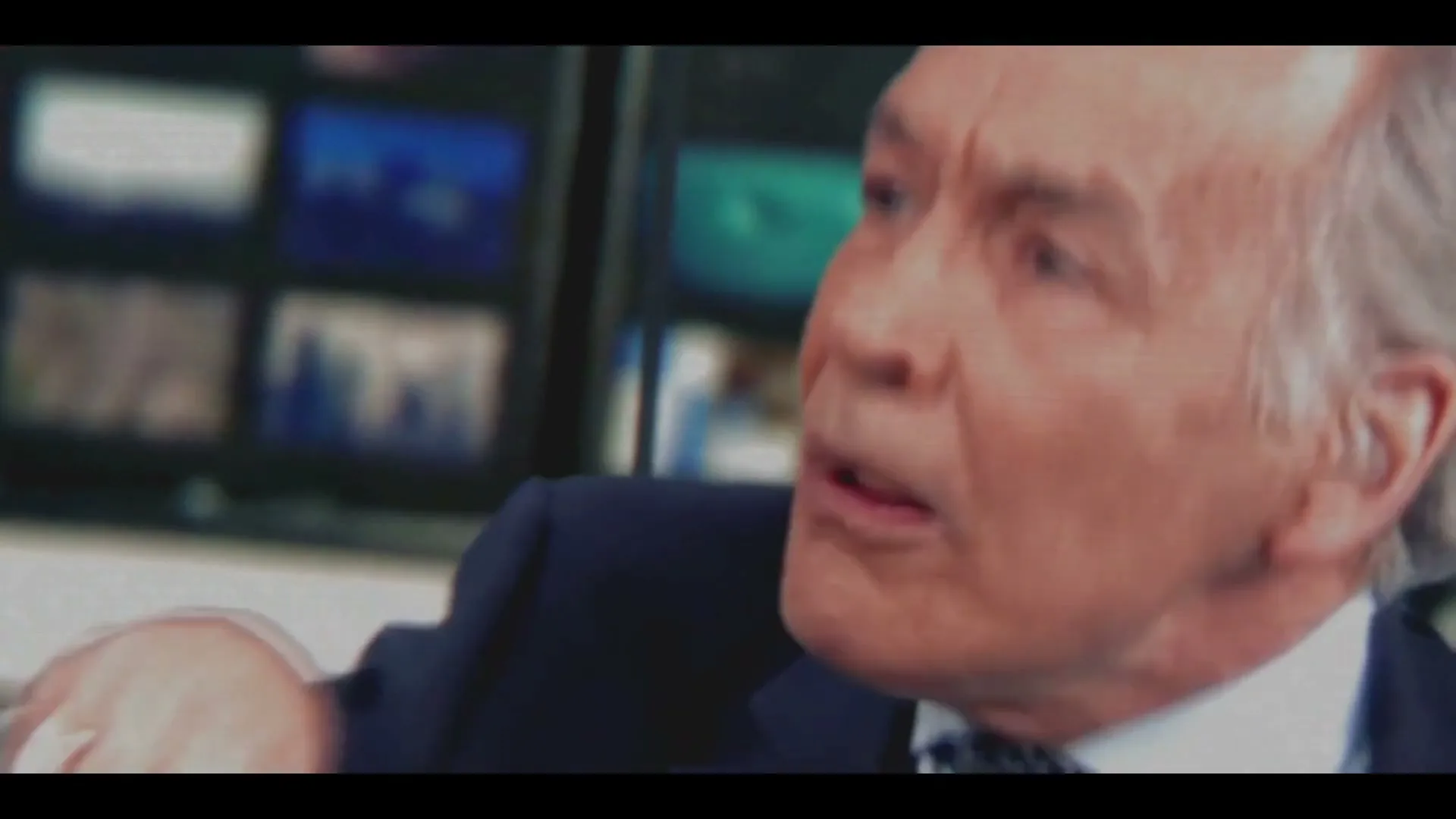 A male news anchor with grey hair and a suit looks concerned while speaking in a news studio with multiple screens in the background.