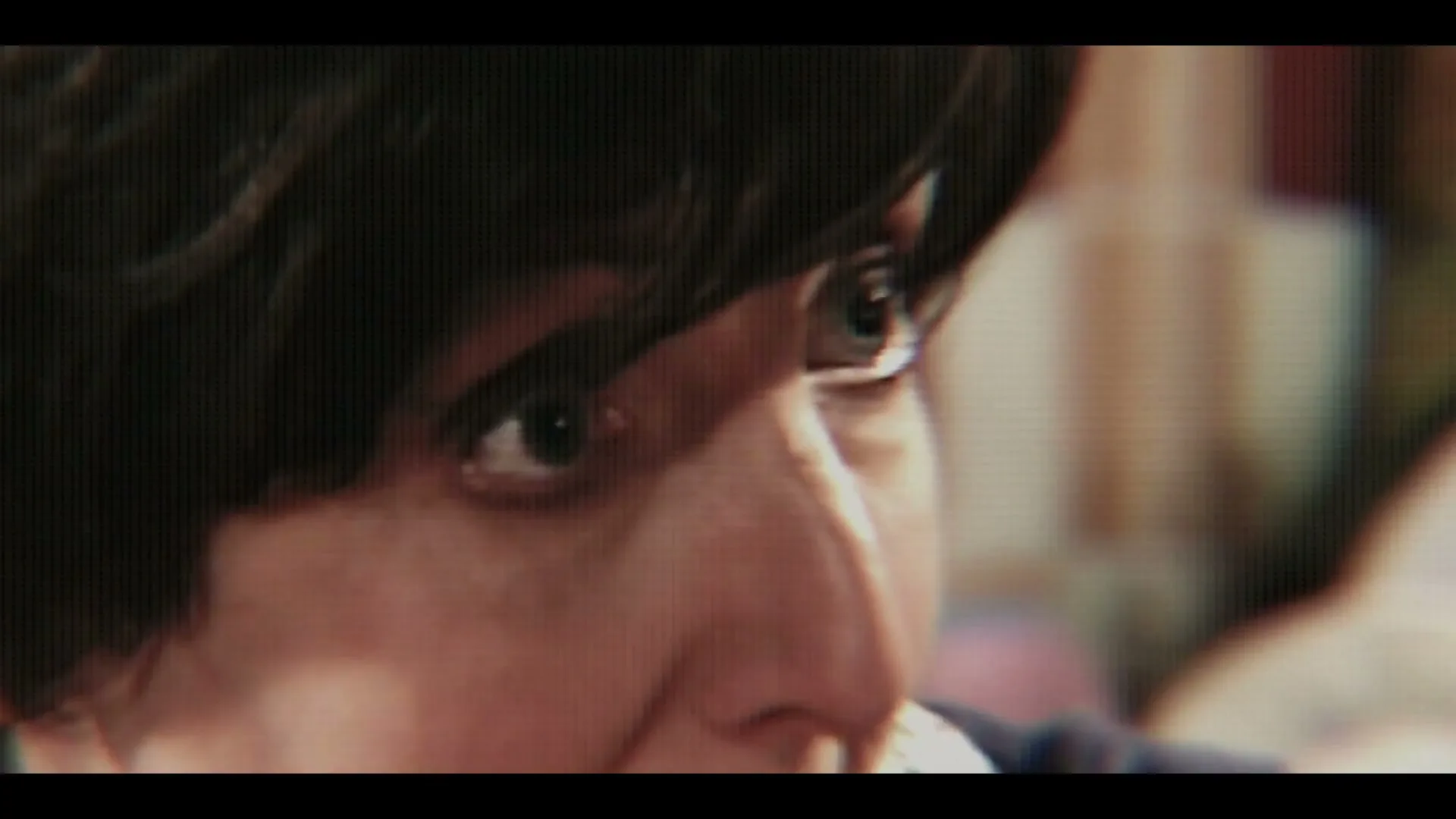 A young person with dark hair and light eyes looks intently to the right, their face partially obscured by a pixelated screen effect, in an interior setting.