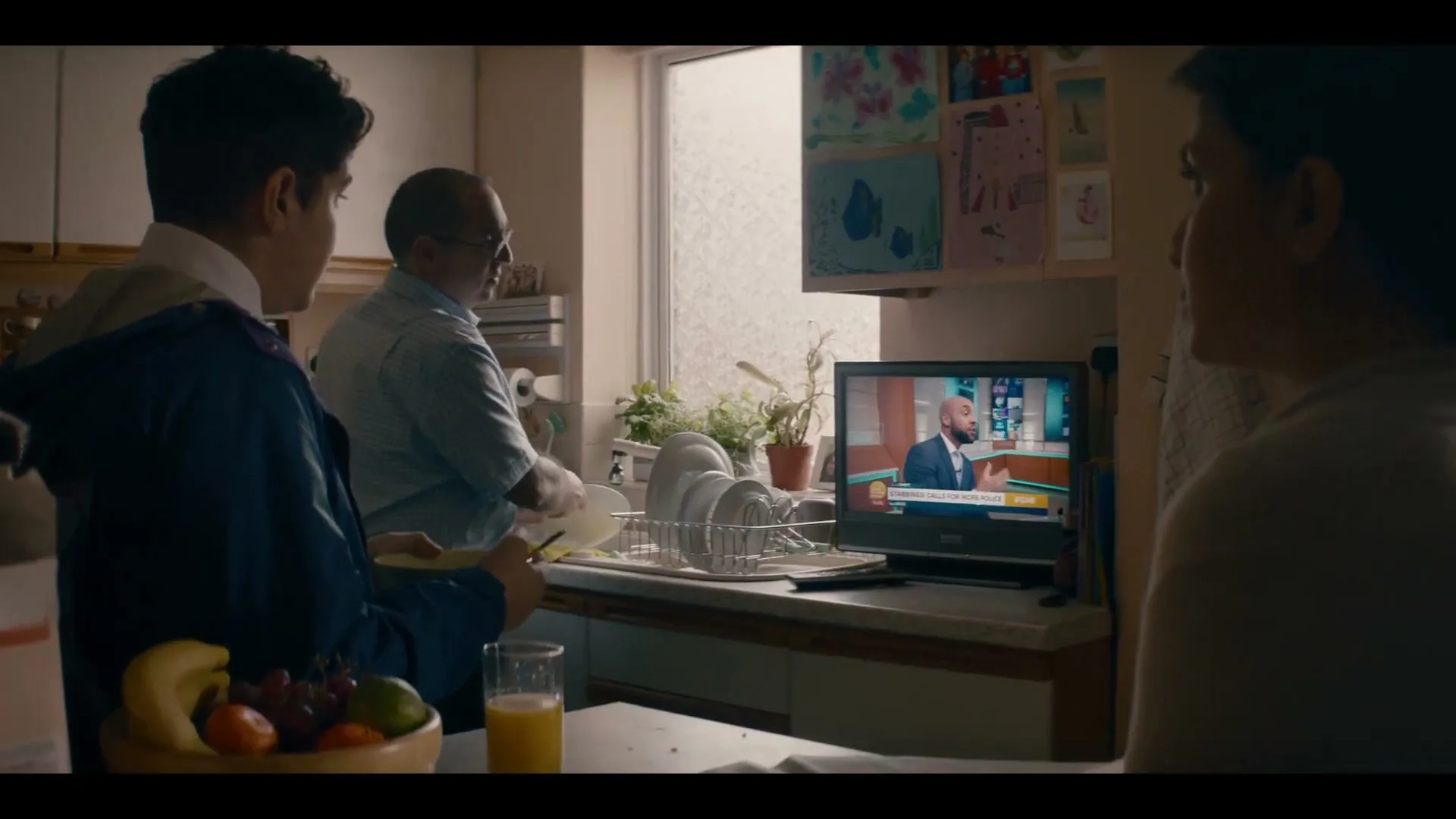 A family of three is in a kitchen, with a man washing dishes, a boy holding a plate, and a woman watching TV.