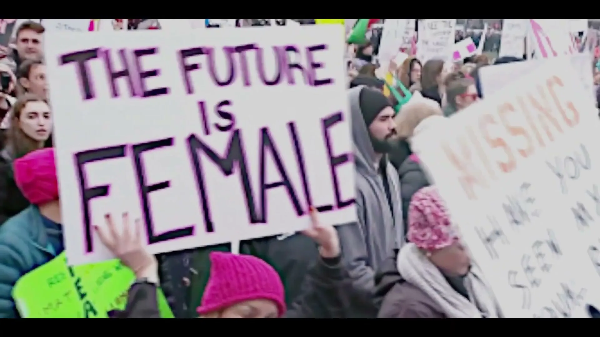 A diverse crowd of people hold up protest signs, including one that reads "THE FUTURE IS FEMALE" and another "MISSING HAVE YOU SEEN MY EQUAL RIGHT" at an outdoor rally.