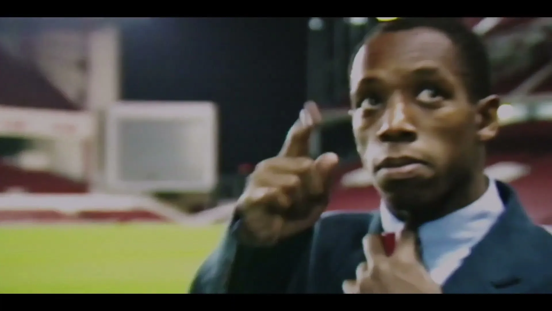 A man in a suit adjusts his tie on a football pitch, looking up with a serious expression as the stadium stands are visible in the background.