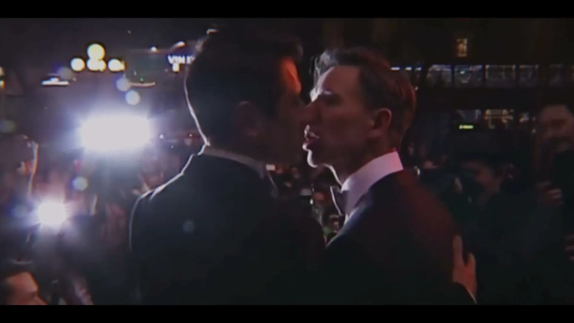 Two men in tuxedos kiss on a red carpet, surrounded by flashing cameras and a crowd of people at night.