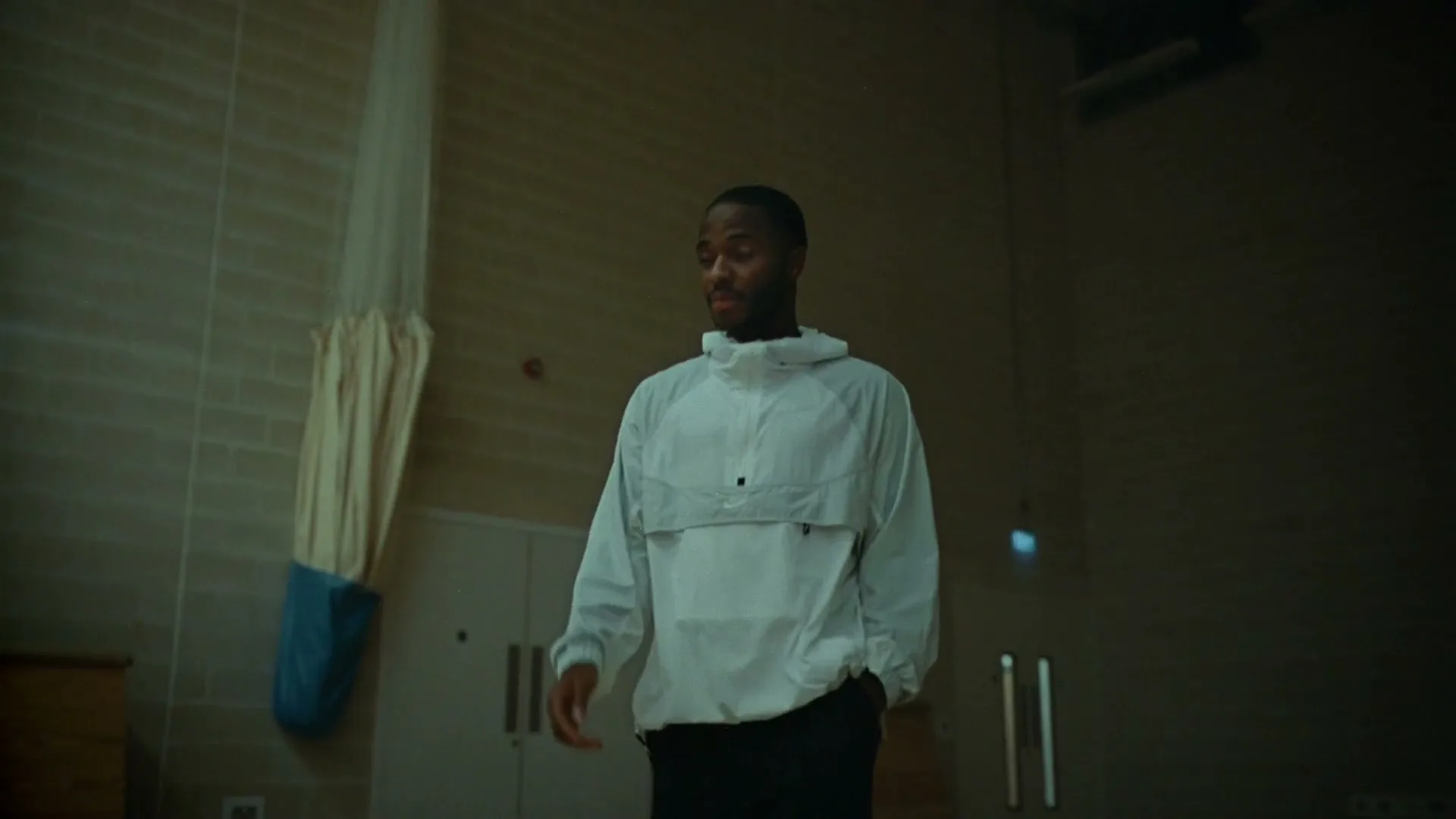 A young Black man in a white windbreaker walks slowly and looks around a dimly lit school gym.