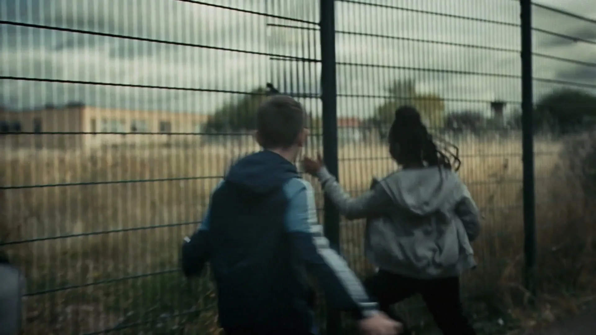 Two children, a boy and a girl, run alongside a chain-link fence, with overgrown grass and buildings visible in the background under a cloudy sky.