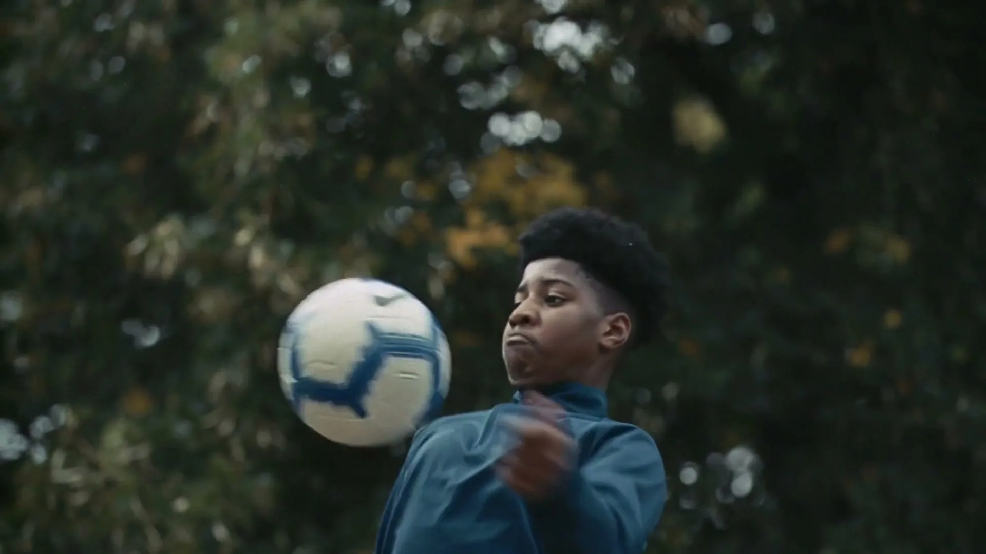 A young Black male in a teal athletic top juggles a soccer ball with his head and shoulder in an outdoor park.