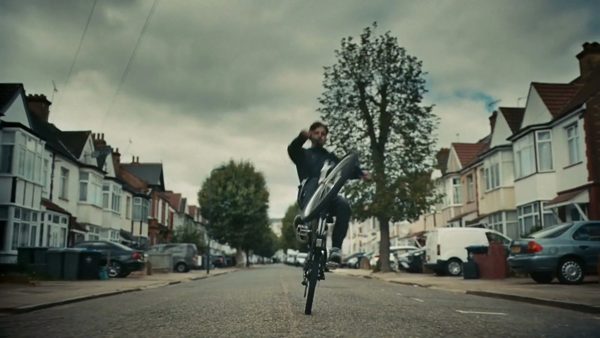 A young man in dark clothing performs a wheelie on a bicycle down a residential street lined with parked cars and houses under a cloudy sky.